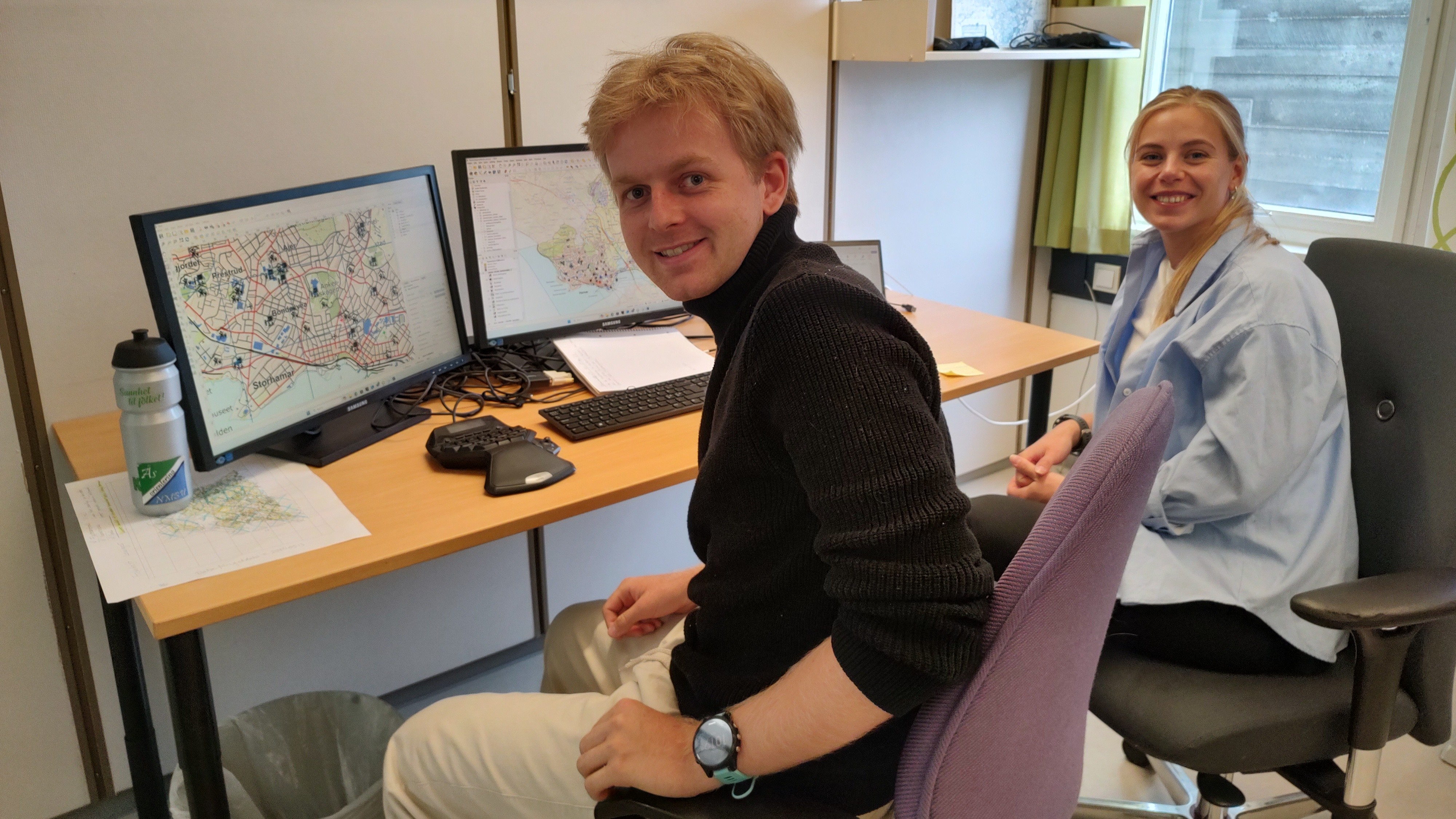 To personer sitter p&aring; et kontor med PC, tastatur m.m. foran seg