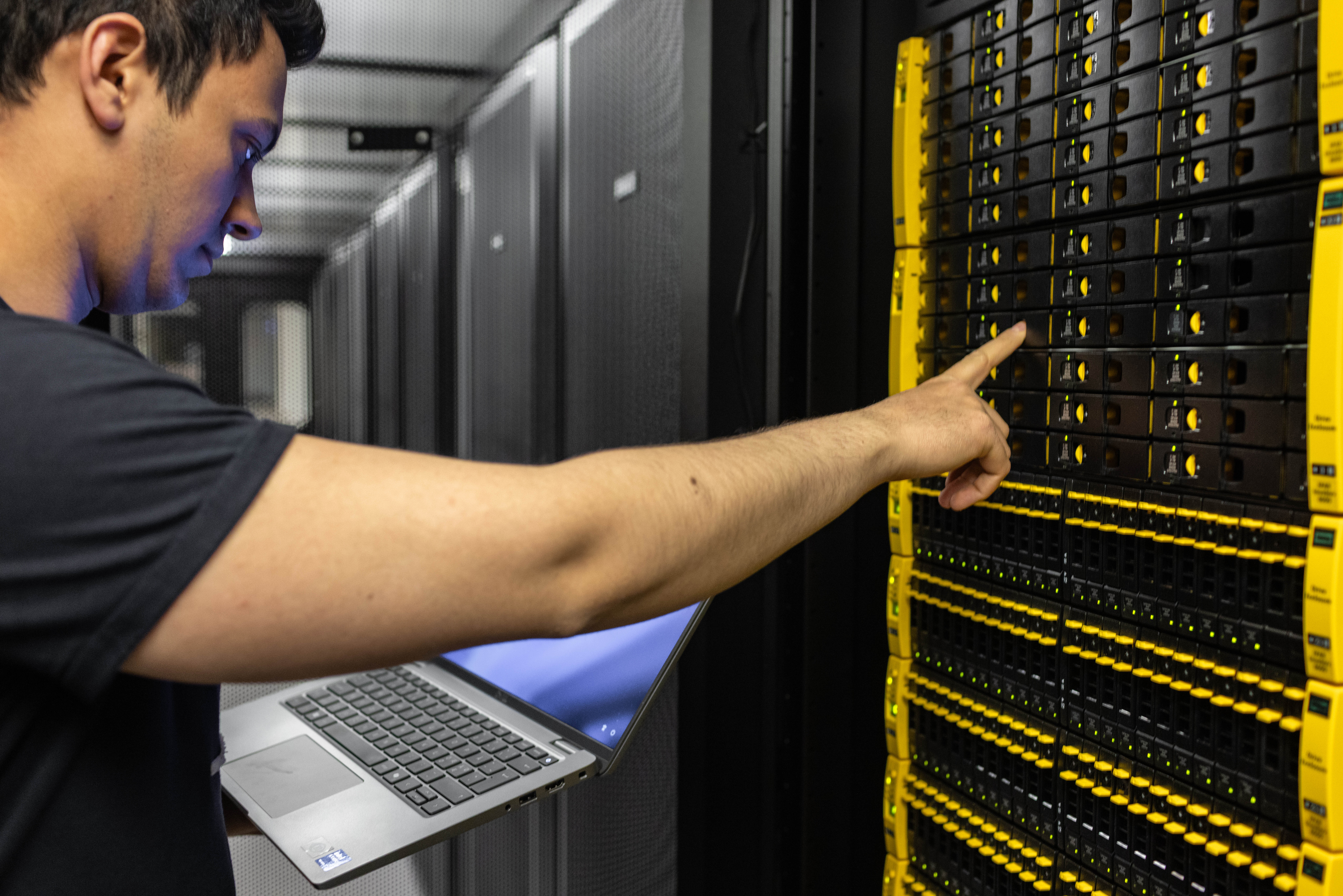 En person står foran et rack med dataservere montert i en reol, mens han ser ned på en bærbar PC.