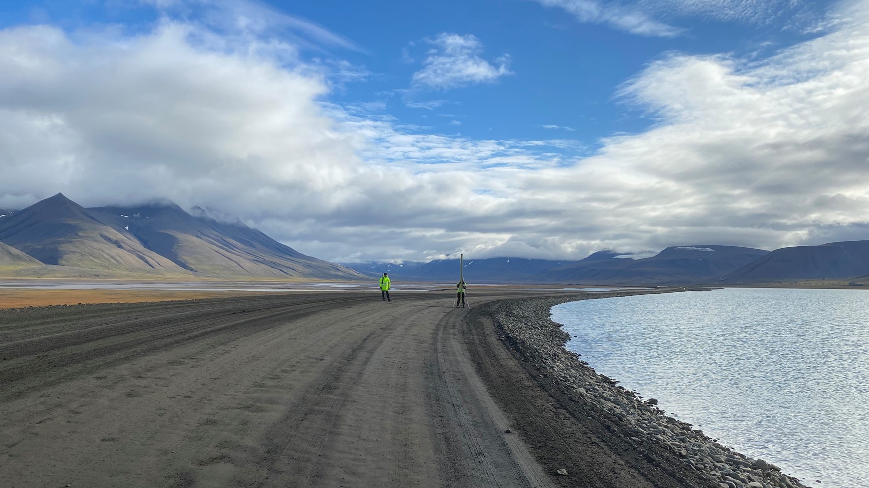 To personer i gule jakker utf&oslash;rer feltarbeid langs en grusvei ved siden av hav, med fjellandskap i bakgrunnen under en skyet himmel.